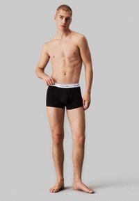 Boxer briefs pretos de algodão com uma cintura de logo branca da Calvin Klein, apresentando um design ajustado e uma textura suave. O modelo está descalço.