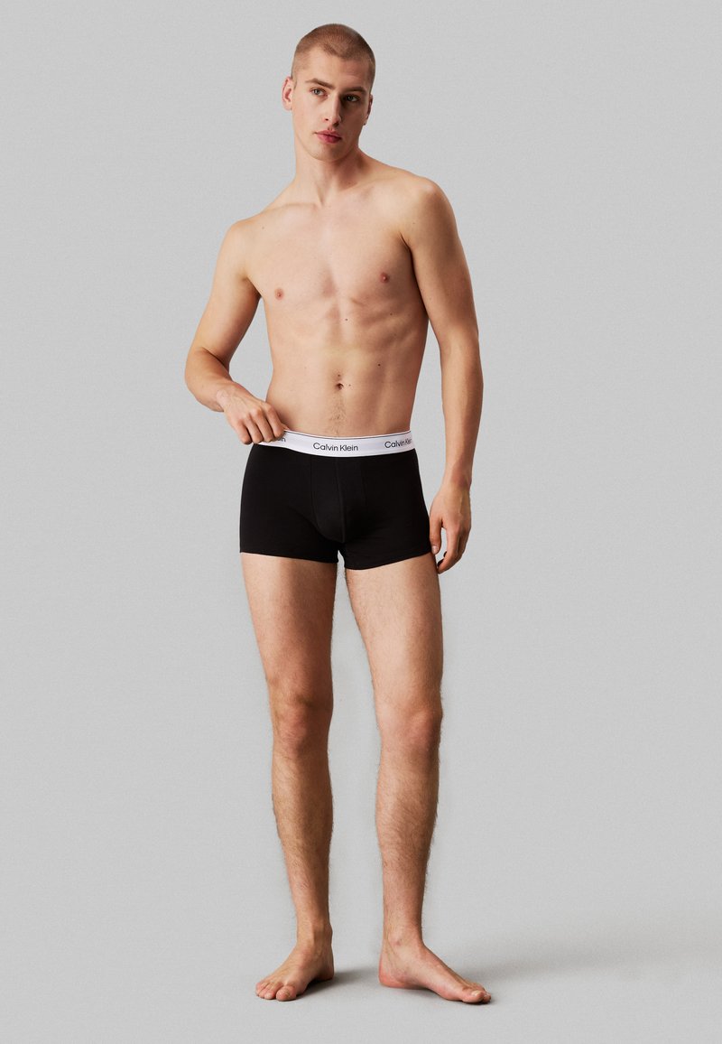 Boxer briefs pretos de algodão com uma cintura de logo branca da Calvin Klein, apresentando um design ajustado e uma textura suave. O modelo está descalço.