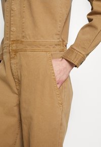 Jumpsuit de algodón marrón con una textura suave, con un cierre frontal abotonado, bolsillos laterales y puños ajustados. Costuras detalladas visibles.
