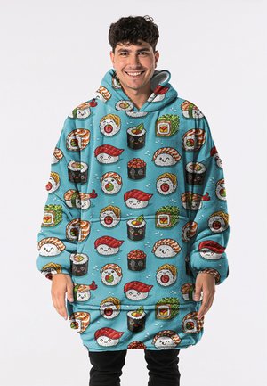 Young man smiling, wearing oversized hoodie with colorful cartoon sushi pieces on bright blue background.