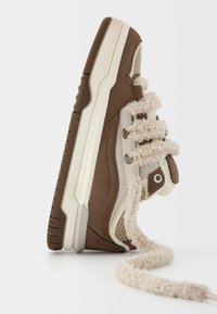 Bruna mockasnedsko med krämfärgade nätpaneler, krämfärgad gummisula och unika fluffiga snören. Har en texturerad tå och en vadderad mellansula.