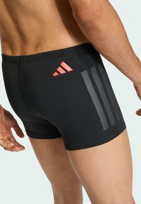 Bañador negro confeccionado en un material elegante, con un logo rojo y tres rayas verticales grises. El diseño incluye un ajuste ceñido y una textura suave.