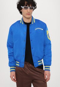 Chaqueta bomber azul con acentos amarillos, botones a presión, y el logo "ICECREAM". Presenta puños acanalados con rayas, llevada sobre una camiseta negra.