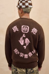 Brauner Strickpullover mit pinkfarbenen grafischen Motiven, darunter Delfine und Text, mit einem gerippten Kragen und Saum. Getragen über Tarnhosen.