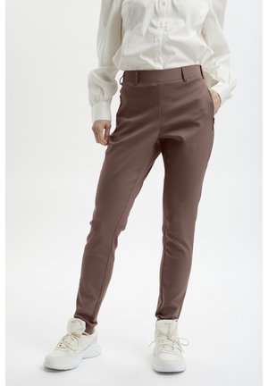 Person, die braune Slim-Fit-Hosen, ein weißes Hemd mit Knöpfen und weiße Sneakers trägt, steht mit einer Hand in der Tasche vor weißem Hintergrund.