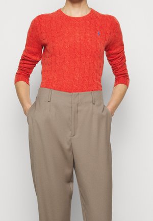 Person iført en rød kabelstrikket sweater stukket ned i beige bukser med høj talje, med hænderne i lommerne mod en ensfarvet baggrund.