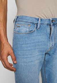 Světle modré džíny s úzkým střihem, které mají klasický design pěti kapes, viditelné švy a značkovou nálepku na zadní kapse.