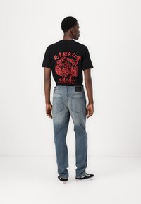 Zwarte t-shirt met rode grafische afbeelding op de achterkant, gecombineerd met vervaagde blauwe denim jeans en zwarte sneakers. Eenvoudig, casual ontwerp.