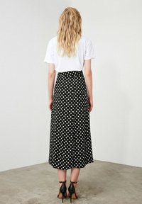 Falda midi negra con lunares, con una cintura ajustada y un diseño fluido, combinada con una camiseta blanca, vista desde la parte de atrás y llevada con tacones negros.