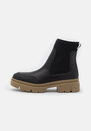 Bottine noire avec semelle en caoutchouc épaisse couleur noisette, panneaux latéraux élastiques et languette à l'arrière, vue de profil sur fond blanc.