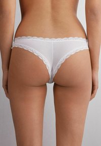 String en dentelle blanche avec un tissu doux et lisse, présentant un délicat bord festonné et une couverture arrière minimale.