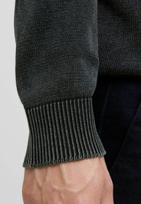 Ciemny wełniany sweter z rękawem eksponującym ściągacz i gładką fakturę, prezentujący mieszankę głębokiej zieleni i szarych włóczek.