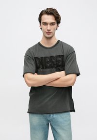 Szürke pamutpóló kerek nyakkivágással és rövid ujjakkal, az elején texturált fekete logóval és nyers szegélyű részletekkel.