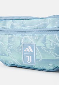 Fanny pack in light blue fabric with abstract patterns; features zip closure and white Adidas logo patch. Textured surface.