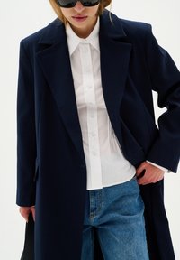 Cappotto oversize blu navy in misto lana, camicia bianca a maniche lunghe e jeans azzurri chiari. Occhiali da sole neri, con vestibilità su misura e rever a punta.