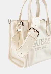 Borsa Guess color crema con due manici, tracolla regolabile con fibbia in argento e testo del brand ricamato sul fronte.