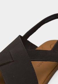 Κοντινή λήψη μαύρου λουριού δερμάτινης σανδάλας με καφέ εσωτερική σόλα, που δείχνει λεπτομερή ραφή και σχεδιασμό με λουράκι στο πίσω μέρος.