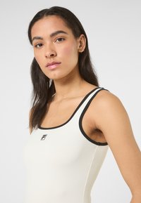Joven con cabello largo y oscuro que lleva una camiseta sin mangas blanca con ribete negro y un pequeño logo en el pecho, mirando al frente con un fondo liso.