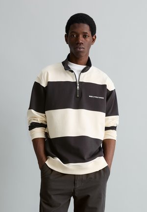 Jonge man die een zwart-wit gestreepte pullover met een kwartrits draagt met de tekst "MARC O'POLO DENIM", handen in de zakken van donkere broeken, neutrale achtergrond.