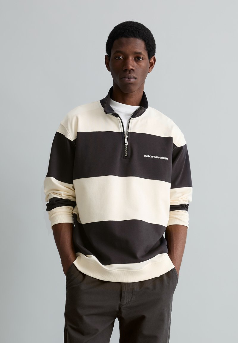 Jeune homme portant un pull à fermeture éclair quart noir et blanc rayé avec le texte "MARC O'POLO DENIM", les mains dans les poches d'un pantalon sombre, fond neutre.