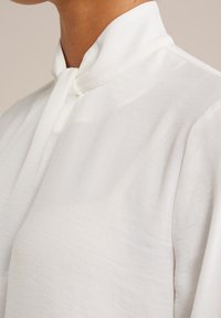 Vit blus med hög krage, mjukt material och knappstängning vid halsen, med lösa, svävande ärmar för en avslappnad passform.