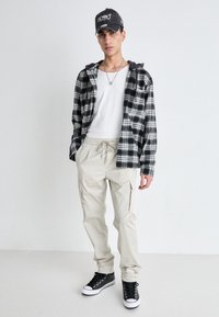 Schwarz-weißer kariertes Flanellhemd mit Kapuze, weißes T-Shirt, beige Cargo-Shorts und schwarze High-Top-Sneaker. Verfügt über eine schwarze Mütze.