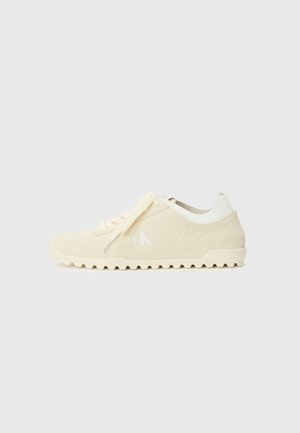Baskets en daim beige avec des accents blancs, semelle texturée et un logo sur le côté. Comprend des lacets plats et un design classique, à profil bas.