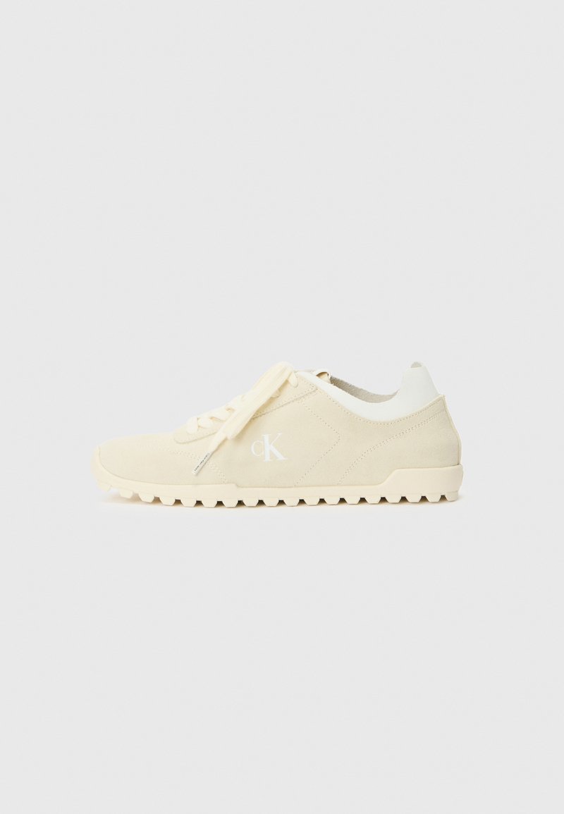 Beige suede sneaker with white accents, textured sole, and a logo on the side. Features flat laces and a classic, low-profile design.