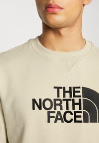 Sudadera beige clara con cuello y puños acanalados. Presenta un logo bordado negro prominente en la parte frontal. Textura suave, diseño casual.