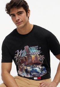 Νέος άνδρας με μαύρο T-shirt που φέρει πολύχρωμο σκηνικό της πόλης του Μαϊάμι, φοίνικες, αγωνιστικό αυτοκίνητο και κείμενο "2026 1-3 Μαΐου" μπροστά.