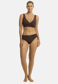 A woman stands confidently, wearing a brown bikini with a wrap-style top and tied bottom, against a plain white background.