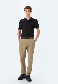 Camisa polo negra con canalé, pantalones beige, cinturón negro y zapatos negros. El modelo está de pie con las manos en los bolsillos frente a un fondo neutro.