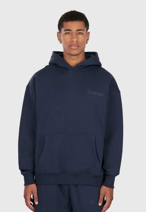 Giovane uomo che indossa una felpa e pantaloni abbinati di colore blu navy con il logo "Dropsize", in piedi contro uno sfondo grigio chiaro uniforme.