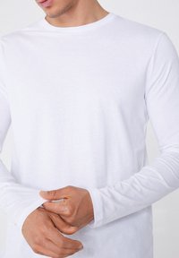 Weißes Langarm-T-Shirt aus weicher Baumwolle. Hat einen runden Ausschnitt und enganliegende Ärmel mit einer glatten, gleichmäßigen Textur. Keine sichtbaren Muster.
