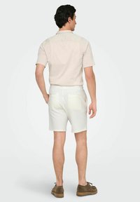 Camisa de manga corta de un color claro combinada con pantalones cortos blancos texturizados y calzado marrón, vista desde atrás. Sin patrones o logotipos destacados.