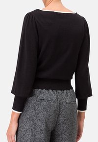 Schwarzer Pullover mit Puffärmeln und weitem Ausschnitt, ausgestattet mit einem kontrastierenden weißen Saum. Getragen mit strukturierten grauen Hahnentritt-Hosen.