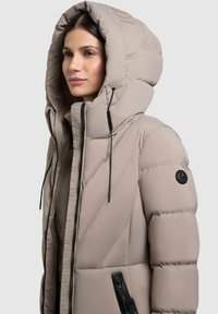 Chaqueta acolchada beige con una capucha grande, costuras diagonales, bolsillos con cremallera y detalles de hardware negro. Textura suave y diseño cálido.