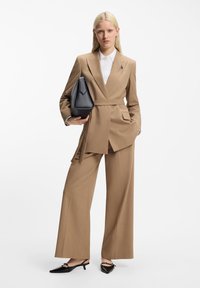 Καφέ κοστούμι με παντελόνι φαρδύ, σακάκι εφαρμοστό και λευκό πουκάμισο. Κρατάει μια μαύρη σκληρή τσάντα, με μαύρα μυτερά παπούτσια.