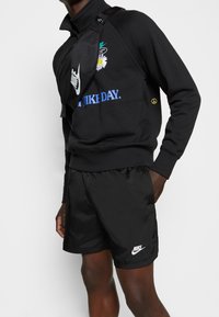 Svart sweatshirt med blommönster och blå text, matchad med svarta shorts med en vit logotyp. Inkluderar en svart axelväska.