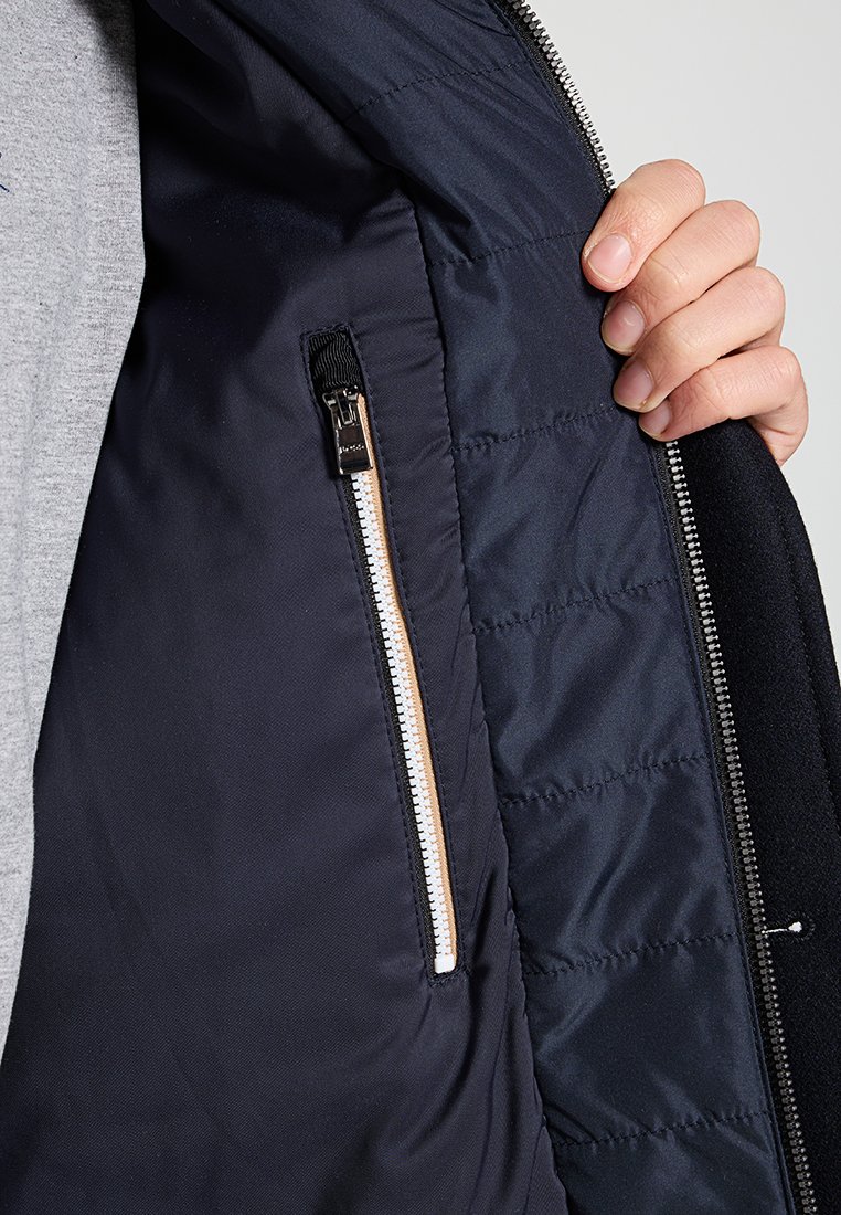 Interno di una giacca blu navale con texture trapuntata, dotata di una tasca con zip in metallo e un accento in pelle marrone. Fodera in tessuto morbido.