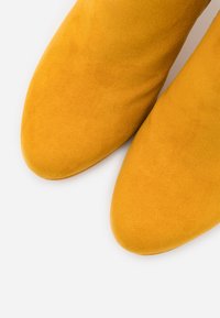 Lähikuva sinapinkeltaisista mokkanahkakenkien varpaista valkoisella taustalla.