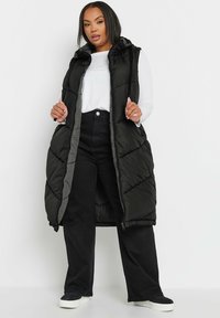 Lång, vadderad svart väst med huva, med en diagonal kviltad mönster och dragkedjestängning. Buren över en vit långärmad topp och svarta byxor.