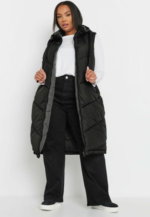 Gilet noir rembourré et long avec capuche, arborant un motif matelassé diagonal et une fermeture éclair. Porté sur un haut à manches longues blanc et un pantalon noir.