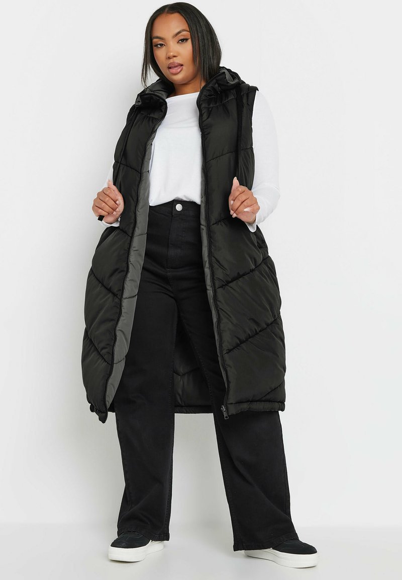 Lång, vadderad svart väst med huva, med en diagonal kviltad mönster och dragkedjestängning. Buren över en vit långärmad topp och svarta byxor.