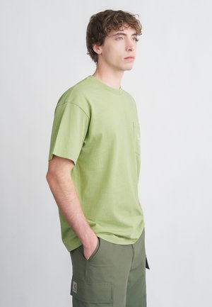 Rövid ujjú, világoszöld pamut póló kerek nyakkivágással és zsebbel, olívazöld cargo nadrággal viselve. Simai felület, laza szabás.