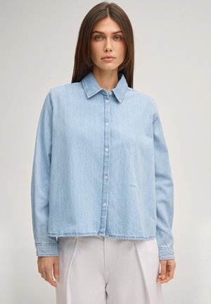 Donna con capelli castani lisci, indossa una camicia di jeans azzurro chiaro con bottoni e pantaloni bianchi, in piedi davanti a uno sfondo semplice.