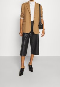 Brauner Blazer, weißer Rollkragenpullover, schwarze Leder-Culottes und schwarze spitze Mules. Kleine schwarze Tasche mit Schultergurt. Durchweg glatte Texturen.