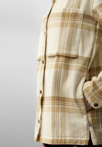 Rutete skjorte i beige og krem med store frontlommer, knappefester og et teksturert stoff. Har en liten detalj ved hemmen.