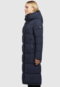 Marineblauwe lange pufferjas met een capuchon, voorzien van een doorgestikt ontwerp, zijzakken en een opvallend logo-patch op de mouw.