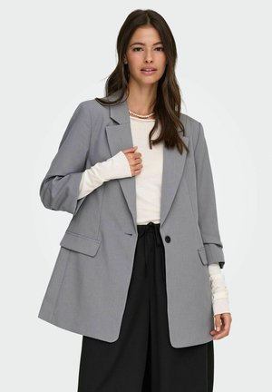 Vrouw die een grijze oversized blazer draagt, een wit overhemd met lange mouwen, zwarte broek en een parelketting tegen een effen lichte achtergrond.